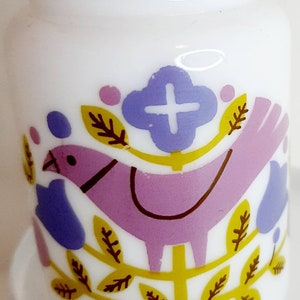 Könnte beinhalten: Weiße Keramikflasche mit einem rosa Vogel, gelben Blättern und blauen Blumen. Der Vogel sitzt auf einem Zweig mit einem weißen Kreuz in der Mitte der Blume.