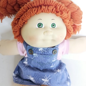 Könnte beinhalten: Eine Puppe mit orangefarbenem Wollhaar in zwei Zöpfen, grünen Augen und heller Haut. Die Puppe trägt ein hellrosa kurzärmeliges Oberteil unter einer blauen Jeanslatzhose mit einem weißen Spinnennetz- und Spinnenmuster sowie zwei braunen Knöpfen.