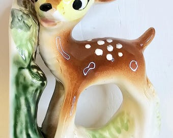 May include: A ceramic figurine of a fawn with brown fur, white spots, and a yellow face. The fawn is standing next to a green and brown tree trunk. The figurine has black hooves and is on a cream-colored base.