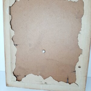 May include: The back of a brown picture frame with a metal hanging hook. The frame has a slightly distressed look with some of the paper backing peeling away.