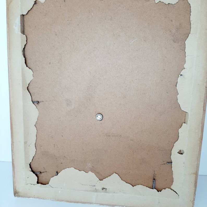 May include: The back of a brown picture frame with a metal hanging hook. The frame has a slightly distressed look with some of the paper backing peeling away.