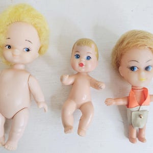 Könnte beinhalten: Drei Vintage-Puppen aus Plastik mit blonden Haaren. Die Puppen sind nackt und haben bemalte Gesichtszüge. Eine Puppe trägt eine kleine orange-braune Weste und einen beigefarbenen Rock.