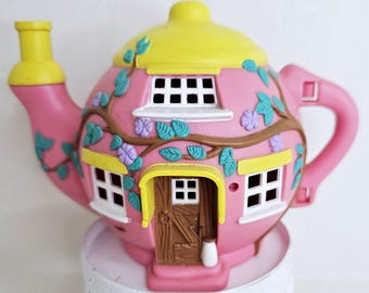 May include: A pink teapot-shaped house with a yellow lid and spout. The house features white-framed windows, a brown door, and decorative vines with blue and purple accents. A whimsical decorative item.