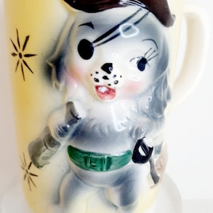Könnte beinhalten: Eine vintage Keramiktasse mit einem Cartoon-Piratenhund-Design. Der Hund trägt einen braunen Hut, eine Augenklappe und einen grünen Gürtel. Die Tasse hat einen gelben Hintergrund mit schwarzen Sternbursts.