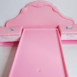 Puede incluir: Un juego de cama de muñeca de plástico rosa con cabecero, estructura de cama y dos mesitas de noche. El cabecero tiene un diseño floral decorativo. Cada mesita de noche tiene un cajón con un diseño floral y una pequeña imagen encima.