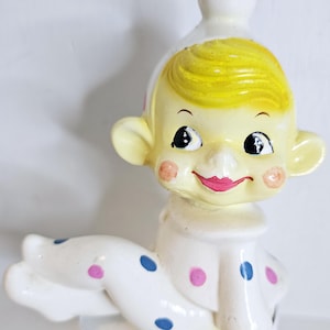 May include: A vintage ceramic figurine of a smiling baby-like figure with yellow hair, rosy cheeks, and a pink mouth. The figure wears a white outfit with blue and pink polka dots. The figurine has large ears and a white top.