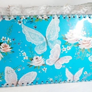 Pu&ograve; includere: Una scatola blu turchese con un bordo di pizzo bianco e un motivo floreale con farfalle e rose. La scatola ha un bordo smerlato.