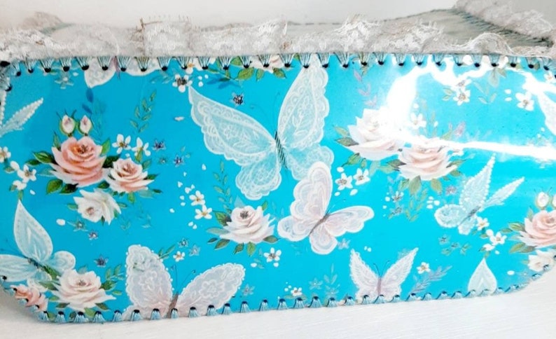 Pu&ograve; includere: Una scatola blu turchese con un bordo di pizzo bianco e un motivo floreale con farfalle e rose. La scatola ha un bordo smerlato.