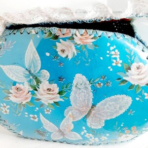 Pu&ograve; includere: Una scatola ovale blu per gioielli con un bordo in pizzo bianco. La scatola &egrave; decorata con un motivo floreale con rose rosa e farfalle bianche.