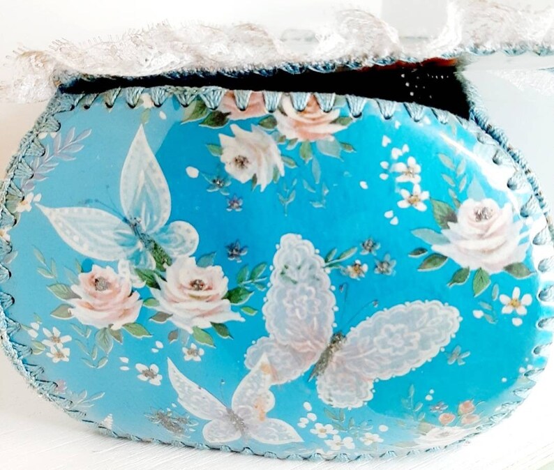 Pu&ograve; includere: Una scatola ovale blu per gioielli con un bordo in pizzo bianco. La scatola &egrave; decorata con un motivo floreale con rose rosa e farfalle bianche.