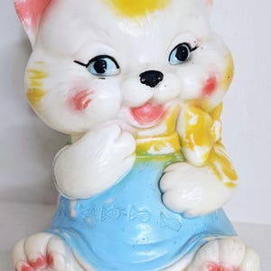 May include: A vintage ceramic cat figurine with a cheerful expression. The cat has white fur, blue eyes, and rosy cheeks. It wears a blue dress with embossed details and yellow accents. The figurine is approximately 6 inches tall.