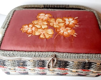 Può includere: Una scatola da cucito vintage in vimini con un motivo floreale sul coperchio. Il coperchio è rivestito in tessuto rosso e presenta fiori gialli ricamati. La scatola ha una chiusura in metallo argentato e un motivo intrecciato sul fondo.