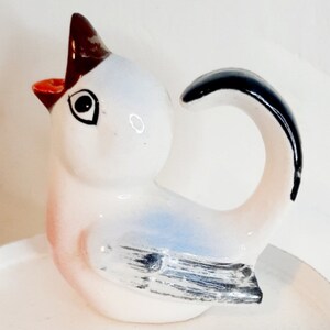 Könnte beinhalten: Eine weiße Keramik-Vogelfigur mit einem blauen und grauen Flügeldesign. Der Vogel hat einen schwarzen Schwanz, der einen Griff bildet. Der Schnabel des Vogels ist offen und rot bemalt.