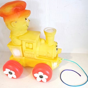 May include: A yellow plastic toy train with a red and orange conductor figure. The train has a pull string and red wheels with white flower designs.