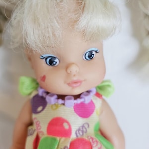 以下が含まれることがあります： ブロンドの髪をツインテールにしたヴィンテージドール。人形は青い目を持ち、カラフルな水玉模様のイエローのドレスと紫のネックレスを着用しています。肩には緑のリボンが付いています。