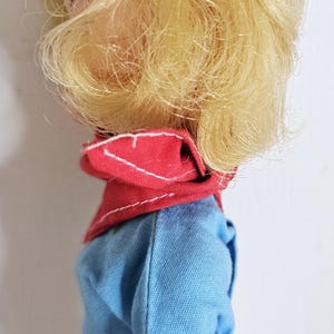K&ouml;nnte beinhalten: Eine Vintage-Puppe mit blondem Haar, das zu einem Pferdeschwanz mit einem wei&szlig;en Band gebunden ist. Die Puppe tr&auml;gt ein blaues Outfit mit rotem Kragen und wei&szlig;em Besatz. Die Haut der Puppe ist hell.