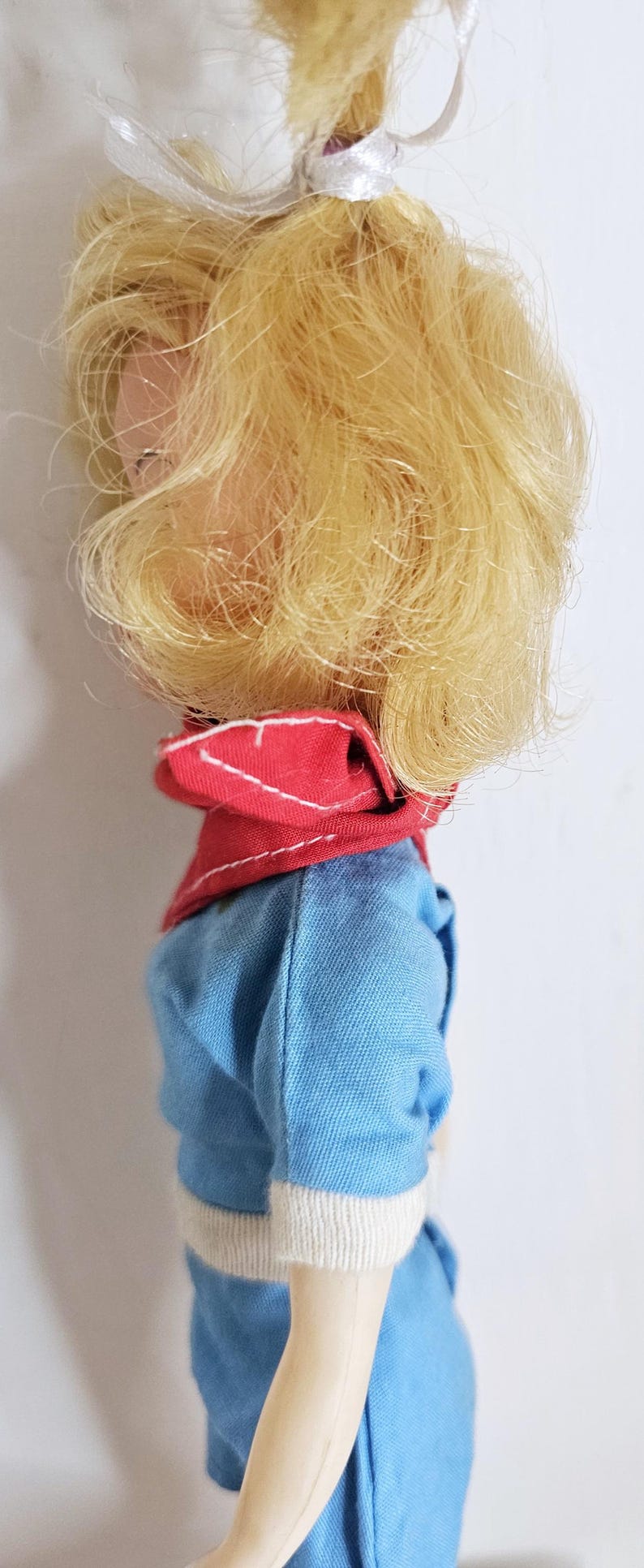 K&ouml;nnte beinhalten: Eine Vintage-Puppe mit blondem Haar, das zu einem Pferdeschwanz mit einem wei&szlig;en Band gebunden ist. Die Puppe tr&auml;gt ein blaues Outfit mit rotem Kragen und wei&szlig;em Besatz. Die Haut der Puppe ist hell.
