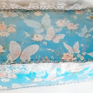 Pu&ograve; includere: Una scatola blu con un bordo di pizzo bianco e un motivo floreale con farfalle e rose. La scatola &egrave; fatta di cartone e ha un aspetto vintage.