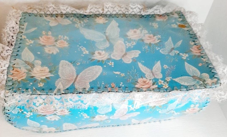 Pu&ograve; includere: Una scatola blu con un bordo di pizzo bianco e un motivo floreale con farfalle e rose. La scatola &egrave; fatta di cartone e ha un aspetto vintage.