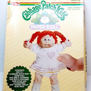 Pode incluir: Um conjunto de bonecas de papel Cabbage Patch Kids com uma boneca de cabelo ruivo, cinco roupas e um bastão de cola. A caixa é amarela com detalhes verdes e brancos e apresenta o logotipo Cabbage Patch Kids.