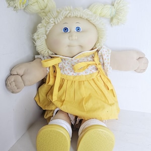 May include: A soft-sculpture doll with pale skin, bright blue eyes, and light yellow yarn hair styled in two high pigtails. The doll wears a white floral-print top with a yellow pinafore dress, white socks, and solid yellow shoes with textured soles.