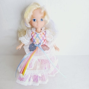 Könnte beinhalten: Eine Puppe mit blondem Haar und blauen Augen trägt ein weißes Kleid mit pastellfarbenem Gittermuster am Oberteil, Spitzenbesatz und einem mehrlagigen Rock. Das Kleid hat zwei glitzernde Blumenapplikationen an der Taille mit langen Bändern.