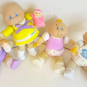 Może przedstawiać: Cztery lalki Cabbage Patch Kids w różnych strojach. Lalki siedzą lub stoją na białej powierzchni. Jedna lalka trzyma małą różową lalkę.