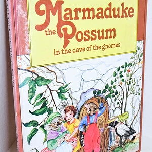Puede incluir: Una portada de libro con fondo amarillo y el título "Marmaduke the Possum in the cave of the gnomes" en texto naranja y marrón. La portada presenta una ilustración de un opossum, un koala, una niña y un cuervo en un entorno de bosque.
