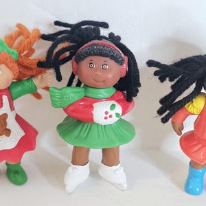 Puede incluir: Tres muñecas pequeñas con diferentes atuendos. Una muñeca viste un atuendo rojo y verde con un delantal de oso, otra un atuendo verde y rojo con una bufanda, y la última un atuendo rojo y amarillo con un sombrero azul.