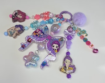 May include: A multi-strand keychain featuring a large purple acrylic bow centerpiece with anime-style character illustrations. It includes various beads in shades of pink, blue, and purple, a fuzzy lavender pom-pom, and letter beads spelling HUNTRIX.