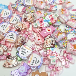 May include: A collection of assorted flatback resin charms featuring various designs including teacups with the text 'have a great day', pink bows, cartoon characters, and small square pillows with colorful patterns in shades of pink, white, and blue.