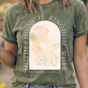 May include: Olive green t-shirt with the text "MOTHERHOOD IS KINGDOM WORK" in a circular design. The shirt features an illustration of a mother and child. The text "PROVERBS 31:25-27" is below the illustration. The Cozy Creative logo is visible.
