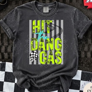 May include: Dark grey t-shirt with the text "Hit the Dang Gas" in neon green and teal. The design includes a checkered flag and tire tracks. Accessories include a checkered hat, sunglasses, and a red koozie.