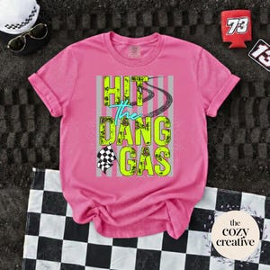 May include: A vibrant pink t-shirt featuring the text "HIT the DANG GAS" in neon green and teal. The design includes tyre tracks and a chequered flag. The shirt is displayed on a black and white chequered surface.