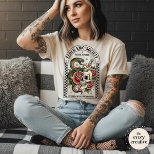 May include: Cream-colored t-shirt featuring a graphic with a skull, snake, and roses, with the text "ELDER EMO SOCIETY NEVER A PHASE". The design is framed by a black and white checkered pattern. The text "EST 1990 METALCORE CRASHOUT" is also included.