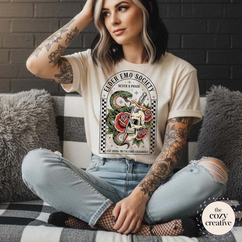 May include: Cream-colored t-shirt featuring a graphic with a skull, snake, and roses, with the text "ELDER EMO SOCIETY NEVER A PHASE". The design is framed by a black and white checkered pattern. The text "EST 1990 METALCORE CRASHOUT" is also included.
