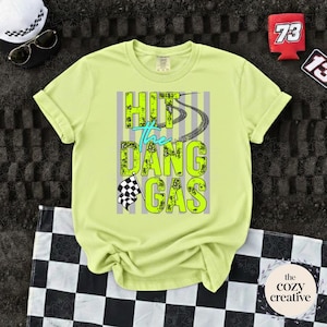 May include: A pale yellow t-shirt featuring the phrase "HIT the DANG GAS" in neon green and teal, with tyre tracks and a chequered flag design. Accessories include sunglasses, a white hat, and a red can cooler.
