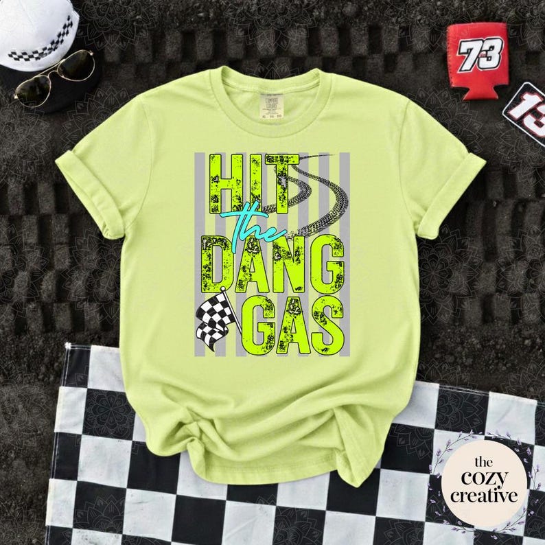 May include: A pale yellow t-shirt featuring the phrase "HIT the DANG GAS" in neon green and teal, with tyre tracks and a chequered flag design. Accessories include sunglasses, a white hat, and a red can cooler.