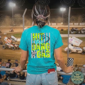May include: Teal short-sleeved t-shirt with the text "Hit the Dang Gas" in neon green and black. The shirt features a checkered flag graphic and a racing track design. The person is at a race track, holding a checkered tumbler.