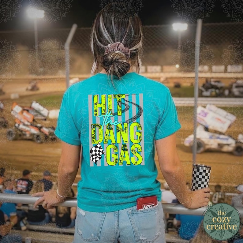 May include: Teal short-sleeved t-shirt with the text "Hit the Dang Gas" in neon green and black. The shirt features a checkered flag graphic and a racing track design. The person is at a race track, holding a checkered tumbler.