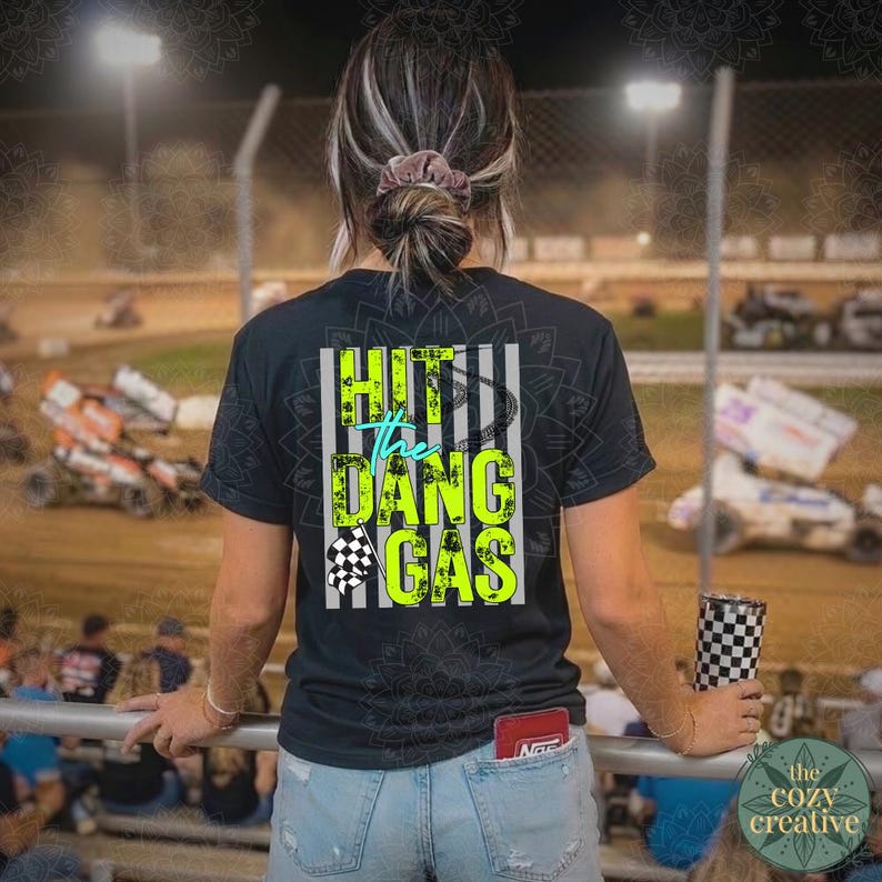 May include: Black t-shirt with the text "HIT THE DANG GAS" in neon green and teal. The shirt features a checkered flag graphic and a vertical striped design. The person is at a race track.