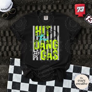 May include: Black t-shirt with the text "HIT the DANG GAS" in neon green and teal. The design includes a checkered flag and a race car track. Accessories include a white hat, sunglasses, and a checkered flag.
