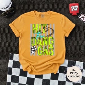 May include: Mustard yellow t-shirt featuring the text "HIT the DANG GAS" in neon green and teal, with tyre tracks and a checkered flag design. The shirt is folded on a black and white checkered surface.