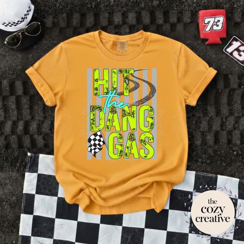May include: Mustard yellow t-shirt featuring the text "HIT the DANG GAS" in neon green and teal, with tyre tracks and a checkered flag design. The shirt is folded on a black and white checkered surface.