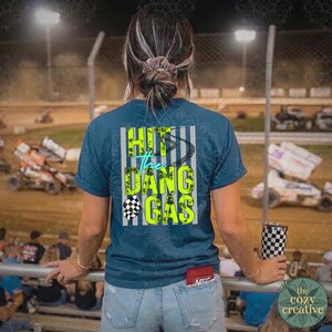May include: A teal t-shirt with the text "Hit the Dang Gas" in neon green and white, and a checkered flag graphic. The person is at a race track, holding a checkered tumbler. The shirt is visible from the back.