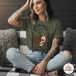 May include: Olive green t-shirt featuring a graphic design with a skull, roses, and a snake. The text on the shirt reads "ELDER EMO SOCIETY NEVER A PHASE" and "EST 1990 METALCORE CHAOS". The shirt is worn with blue jeans.