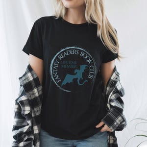 May include: Black t-shirt with a circular design featuring the text "FANTASY READERS BOOK CLUB" and a silhouette of a dragon. The words "LIFETIME MEMBER" are also included in the design. The shirt is worn with a black and white plaid shirt.