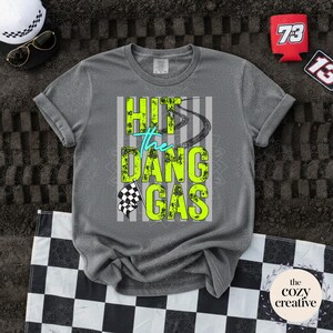 May include: Grey t-shirt featuring the neon green and teal text "HIT the DANG GAS". The design includes a tyre track and a chequered flag. A black and white chequered hat and sunglasses are visible.