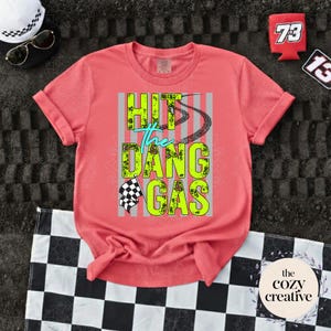 May include: A coral-coloured t-shirt with the text "HIT the DANG GAS" in neon green and teal. The design includes tyre tracks and a chequered flag. Accessories include a white hat, sunglasses, and a racing-themed can cooler.