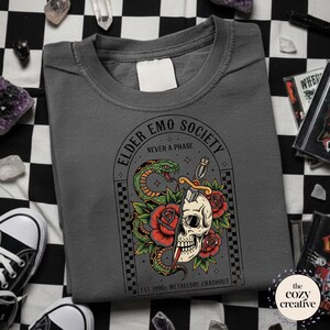 May include: Dark gray t-shirt with a graphic featuring a skull pierced by a dagger, a green snake, and red roses. The text reads "ELDER EMO SOCIETY NEVER A PHASE EST 1990s METALCORE CRASHOUT".